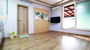 A room with wooden flooring, a wooden door, a wall-mounted TV, and a window with a red roof visible through it.