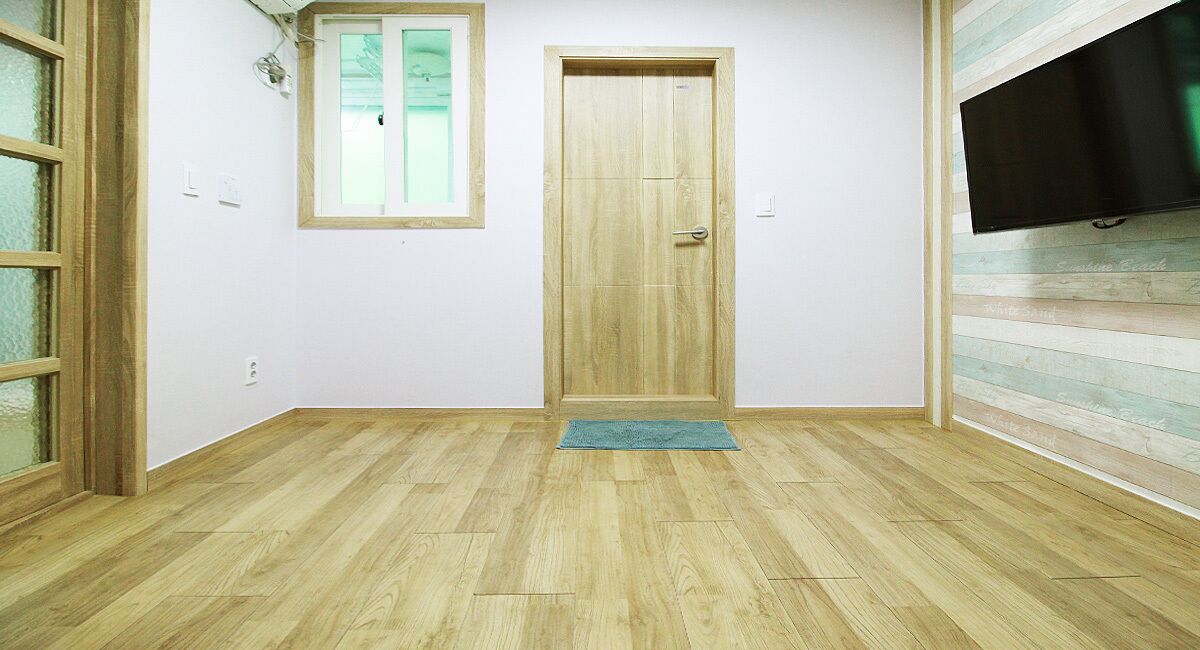 Ein Zimmer mit Holzboden, einer Tür, einem Fenster und einem an der Wand befestigten Fernseher.