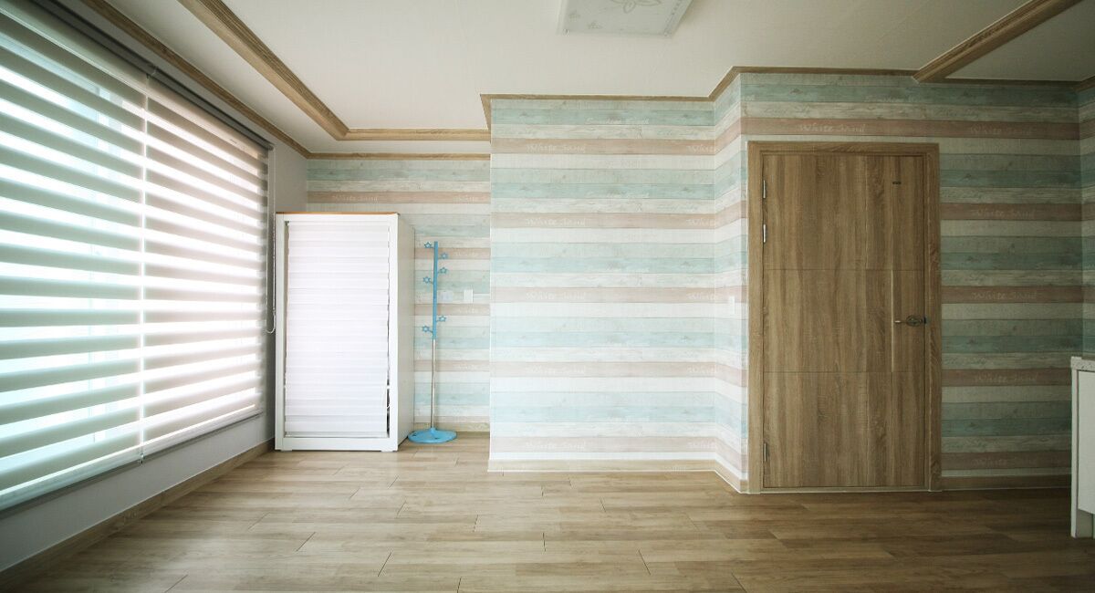 A room with wooden flooring, a large window with blinds, a closed door, and a striped wall.