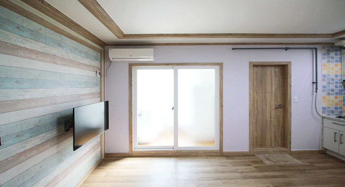 Una habitación con piso de madera, un televisor de pantalla plana montado en la pared, una puerta corrediza de vidrio y un salpicadero de azulejos en la zona de la cocina.