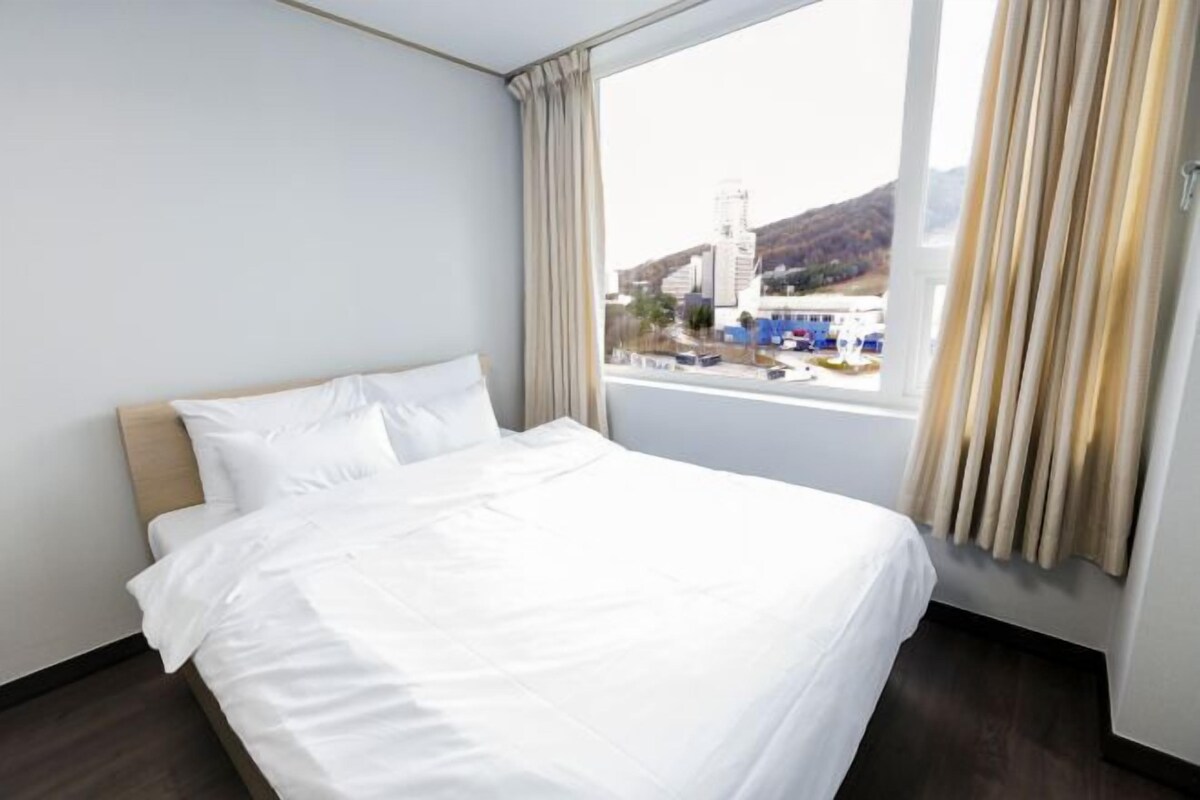 A hotel room with a large bed, a view of a cityscape through a window, and curtains on the window.