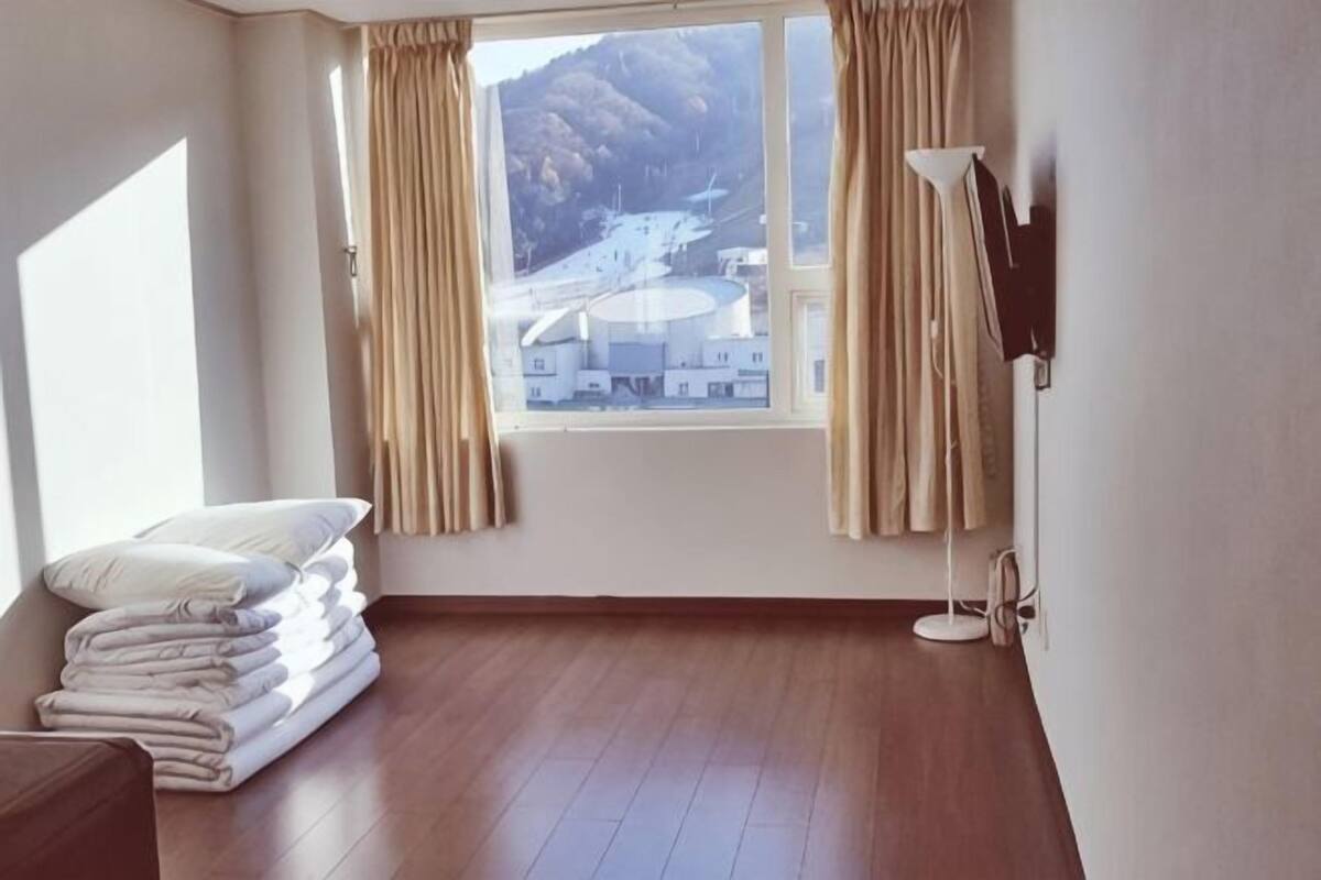 A room with a wooden floor, a stack of white towels, a window with curtains, and a lamp.