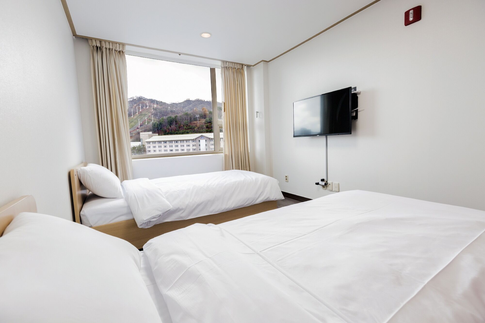 A hotel room with two beds, a TV on the wall, and a window with a view of buildings and hills.