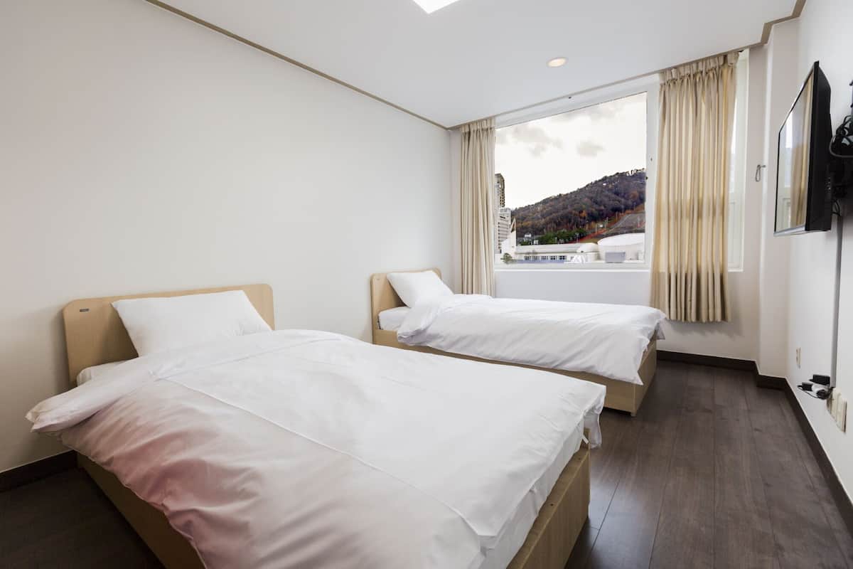 A hotel room with two beds, a large window with curtains, a flat-screen TV mounted on the wall, and a view of a cityscape and hills.