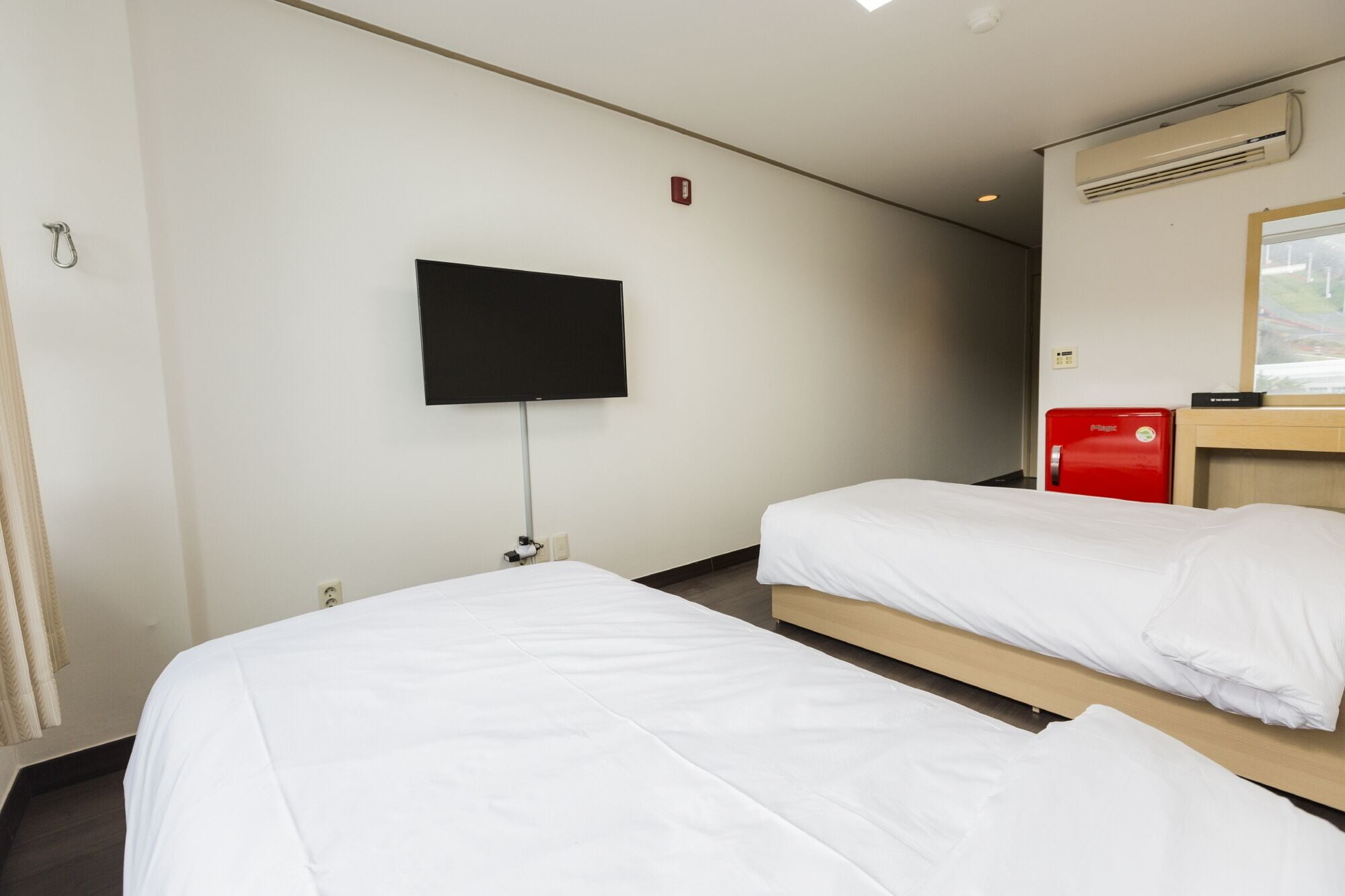 A hotel room with two single beds, a flat-screen TV mounted on the wall, an air conditioning unit, and a red minibar.