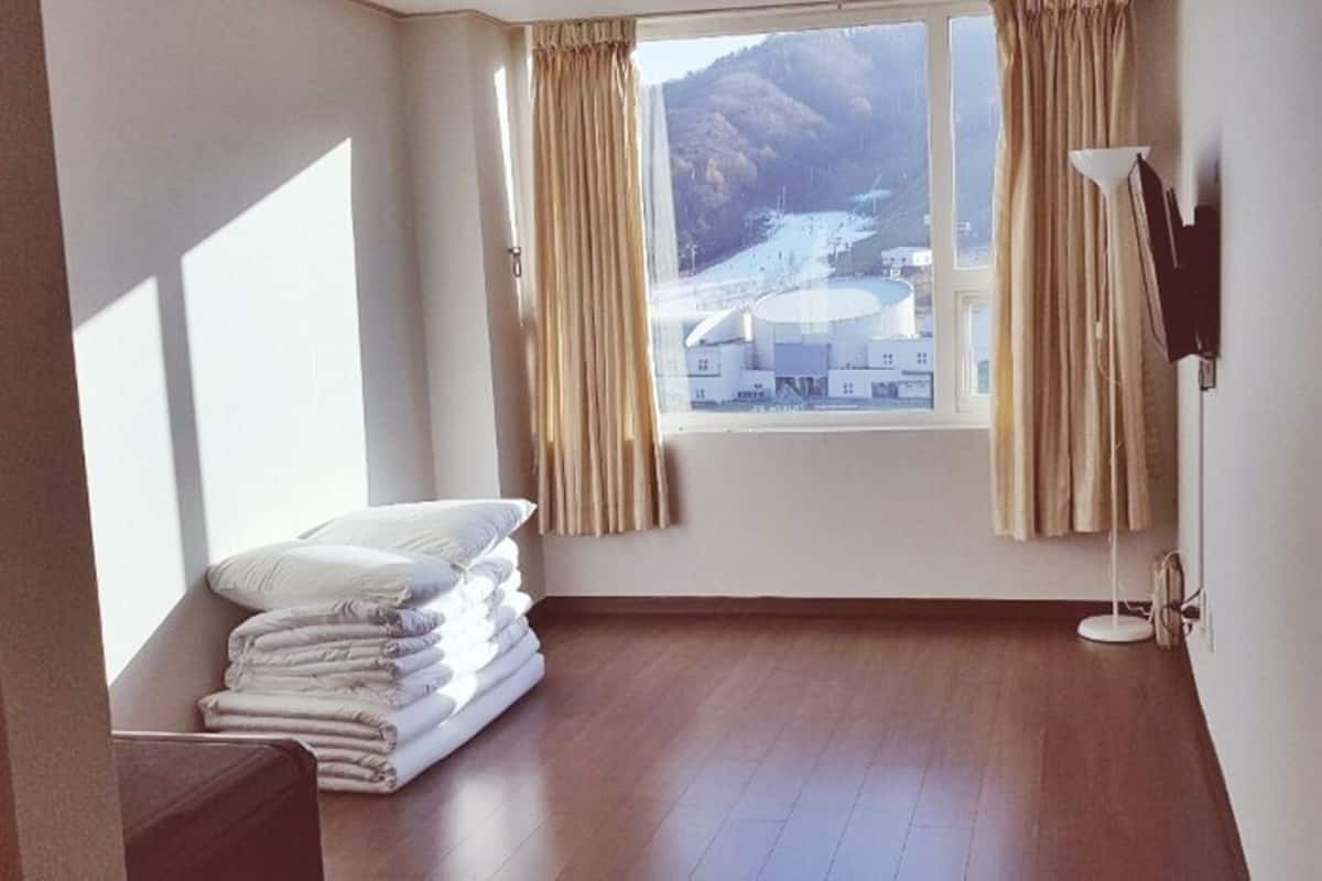 A room with a wooden floor, a bed with white linens, a window with curtains, and a lamp.