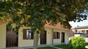 Un edificio con fachada amarilla, varias ventanas con contraventanas marrones y un pequeño jardín con un árbol y un arbusto.