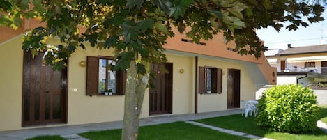 A building with a yellow facade, multiple windows with brown shutters, and a small garden area with a tree and a bush.