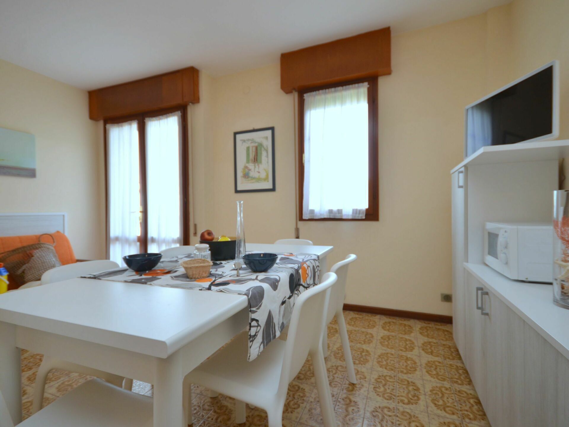 A dining area with a white table, chairs, and a patterned tablecloth. There is a microwave on a white cabinet to the right.