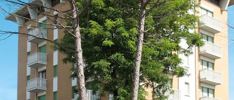 A multi-story building with balconies, a parking area, and a small tree in the foreground.