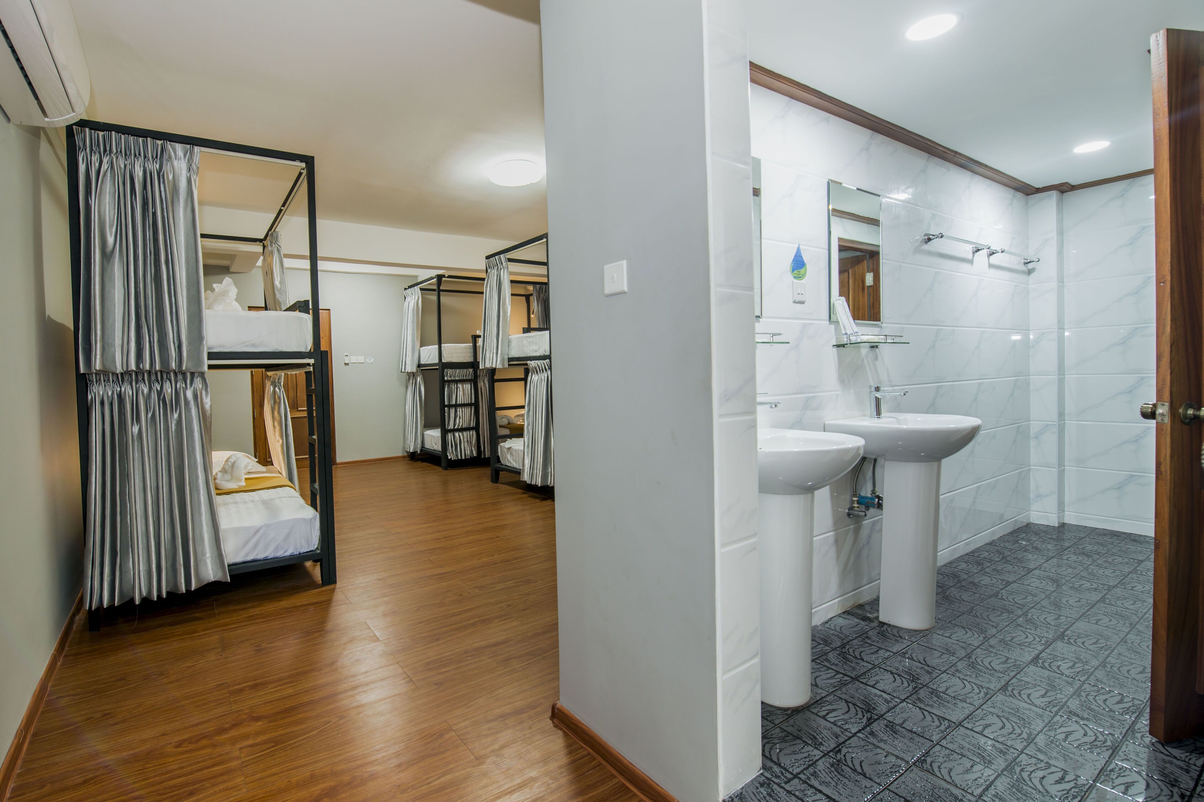 Habitación de hotel con una cama litera, un baño con lavamanos y espejo, y piso de madera.