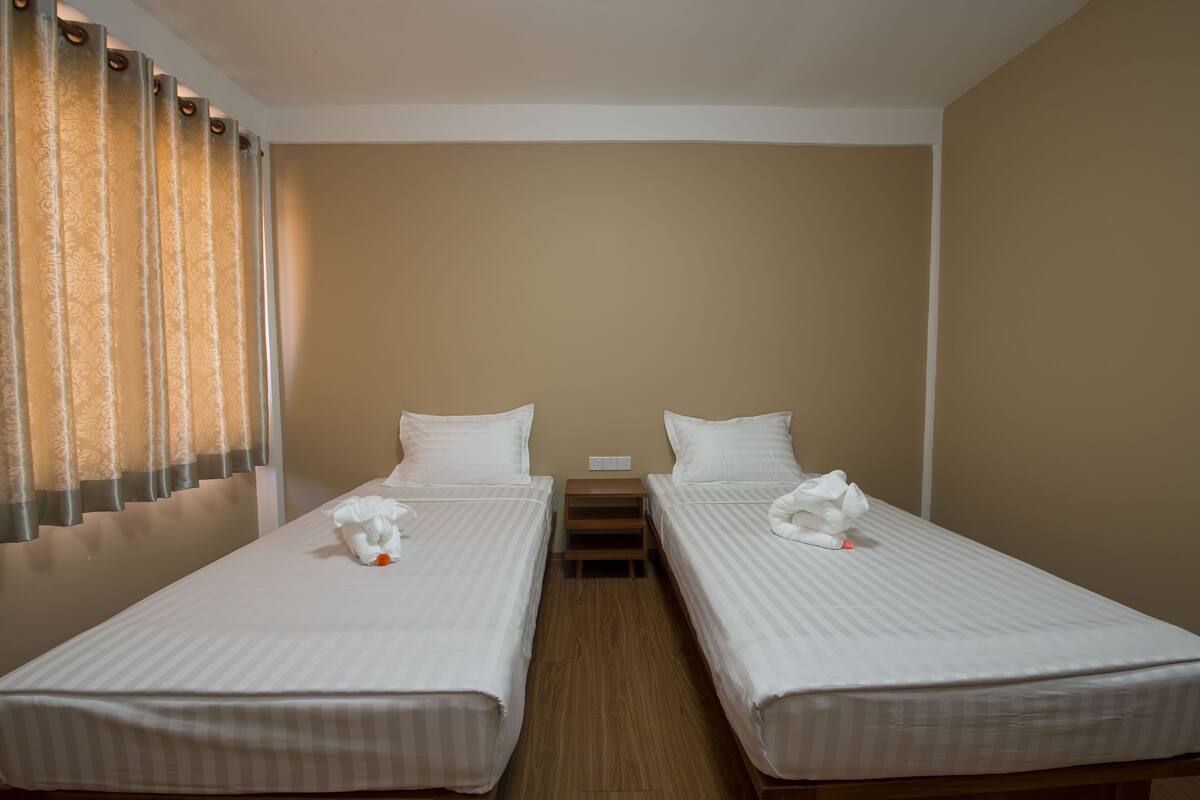 A hotel room with two single beds, a wooden nightstand between them, and a window with curtains.
