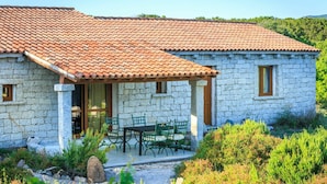 Ein Steinhaus mit Ziegeldach, einer überdachten Terrasse mit Gartenmöbeln und einem Garten mit verschiedenen Pflanzen.