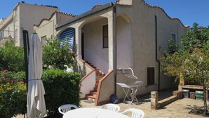 A building with a patio area featuring a round table and chairs, a staircase, and a small garden with plants and a tree.
