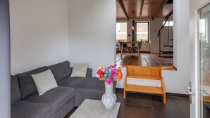 A modern living room with a gray sofa, a wooden bench, and a vase of colorful flowers. There is a dining area with a table and chairs in the background. - Wonderful private villa for 5 people with WIFI, TV, terrace and pets allowed (Schoondijke)