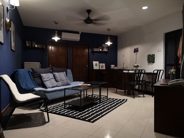 A modern living room with a sofa, coffee table, and dining area. There are ceiling fans, wall-mounted lights, and a large window. - Aurora Court (George Town)