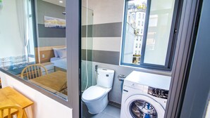 A compact bathroom with a toilet, washing machine, and a glass shower enclosure. - The Art - An Homestay (Da Nang)
