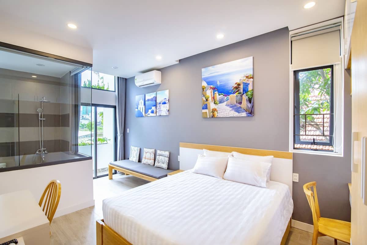 A modern hotel room with a large bed, a seating area with a bench, and a view of the outdoors through large windows.