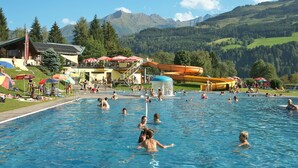 A swimming pool with people, a water slide, and mountains in the background. - Beautiful  villa for 16 guests with WIFI, TV and terrace (Piesendorf)