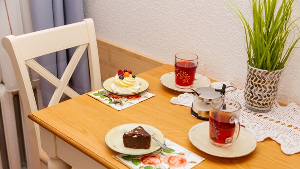 Una mesa de madera con una silla blanca, un plato con una porción de pastel, un vaso de bebida roja y una planta en maceta.