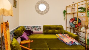 Un sofá verde con una manta de diseño, una silla de madera, un espejo redondo y una estantería con objetos decorativos.