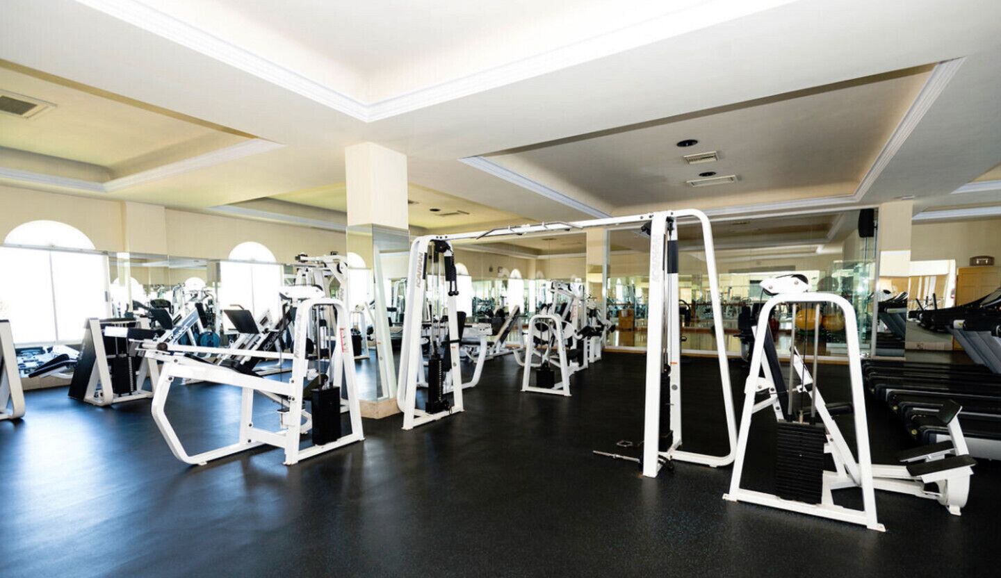 A gym with various exercise machines, large windows, and a polished floor.