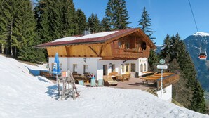 A wooden chalet with a red roof, outdoor seating, and a mountain backdrop.