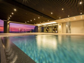 Une piscine intérieure moderne offrant une vue sur le coucher de soleil, dotée d’un éclairage contemporain et de fauteuils suspendus.