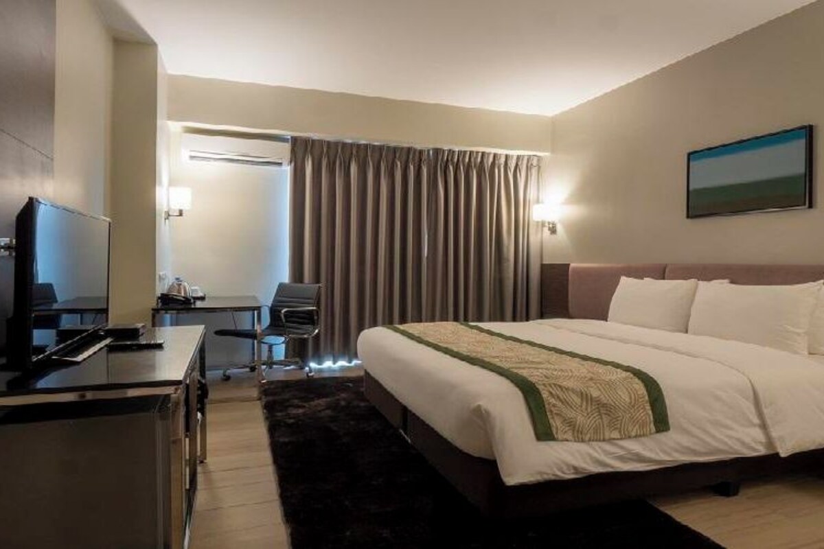 A hotel room with a bed, a desk with a computer, a chair, a TV, and a window with curtains.