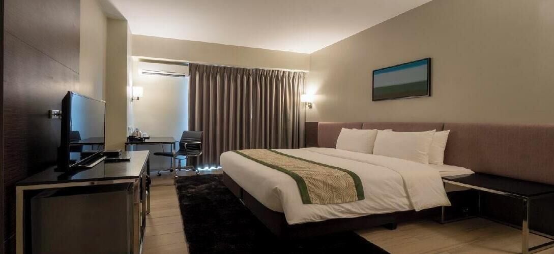 Une chambre d’hôtel avec un lit, un bureau avec un ordinateur, une chaise, une télévision et une fenêtre avec des rideaux.