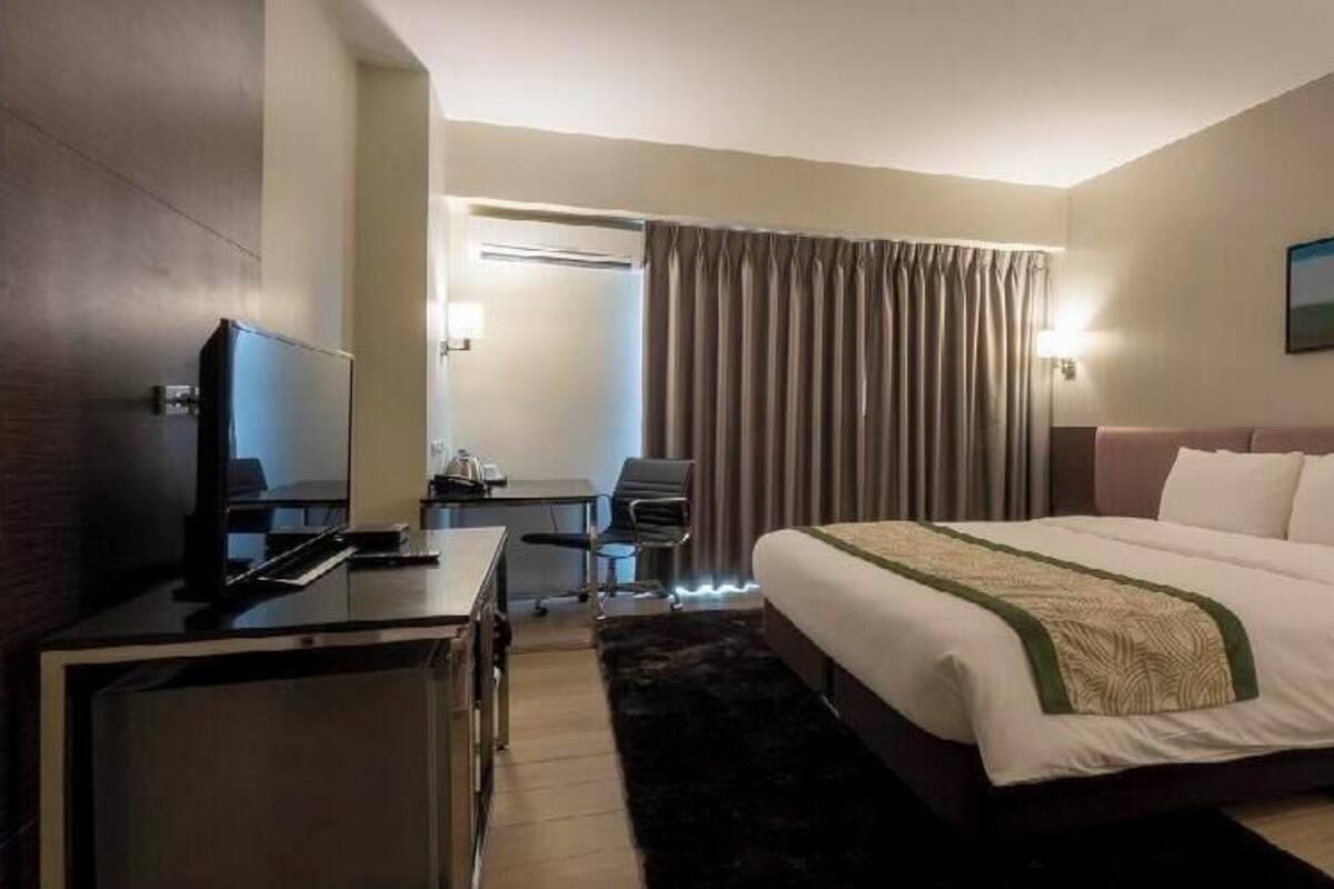 A hotel room with a bed, a desk with a computer, a chair, a TV, and a window with curtains.