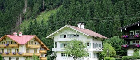 Un bâtiment de style chalet traditionnel, avec un toit rouge, entouré d’une végétation luxuriante et situé au pied d’une colline boisée.