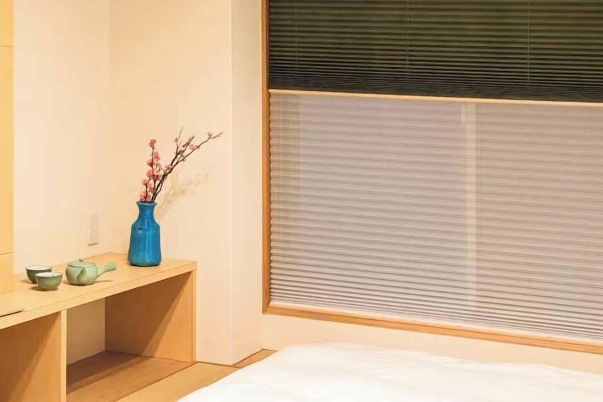 A bedroom with a bed, a bedside table with a vase and teapot, and a window with blinds.