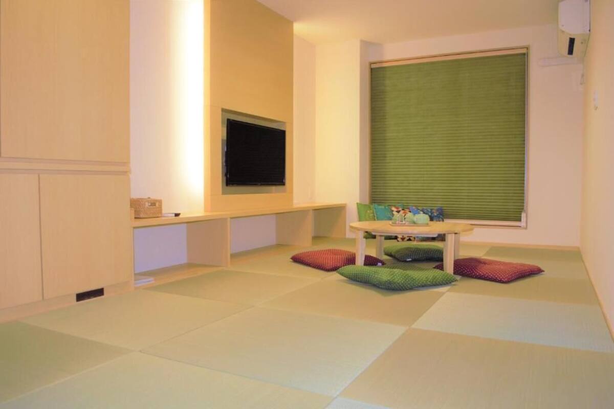 A room with a television mounted on the wall, a small table with cushions, and a window with blinds.