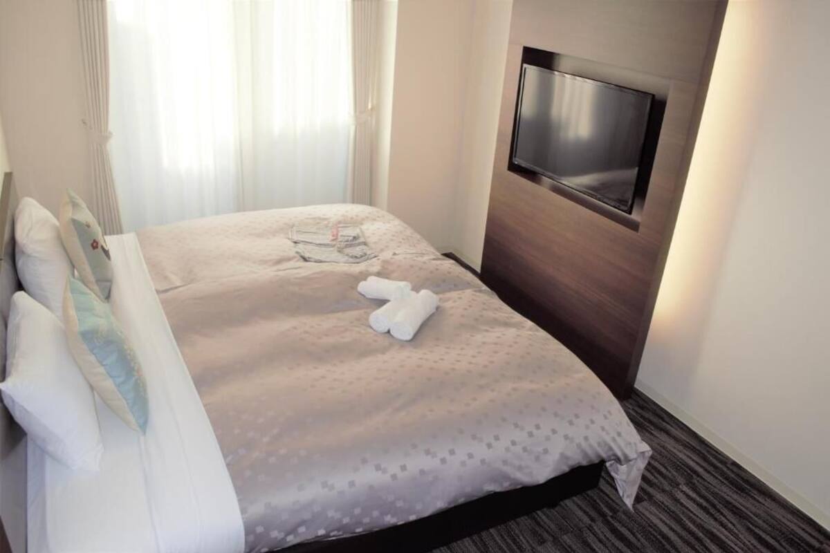 A neatly made bed with a patterned bedspread, a flat-screen TV mounted on the wall, and a window with sheer curtains.