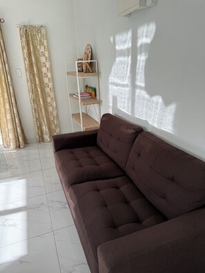 A living room with a brown sofa, a white tiled floor, a white wall, and a window casting shadows. - Moeroa Villa - In the heart of Town. (Avarua)