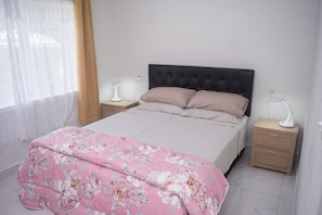 Un dormitorio con una cama, dos mesitas de noche, un enchufe y una ventana con cortinas.