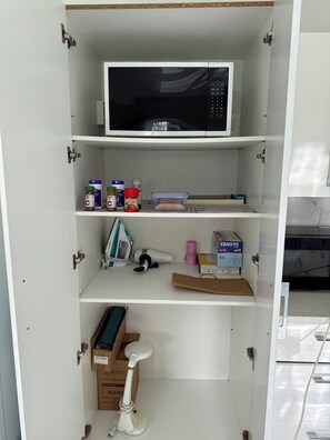 Ein weißer Schrank mit einer Mikrowelle auf der obersten Ablage, verschiedenen Küchenutensilien in der mittleren Ablage und einem weißen Elektroroller auf der untersten Ablage.