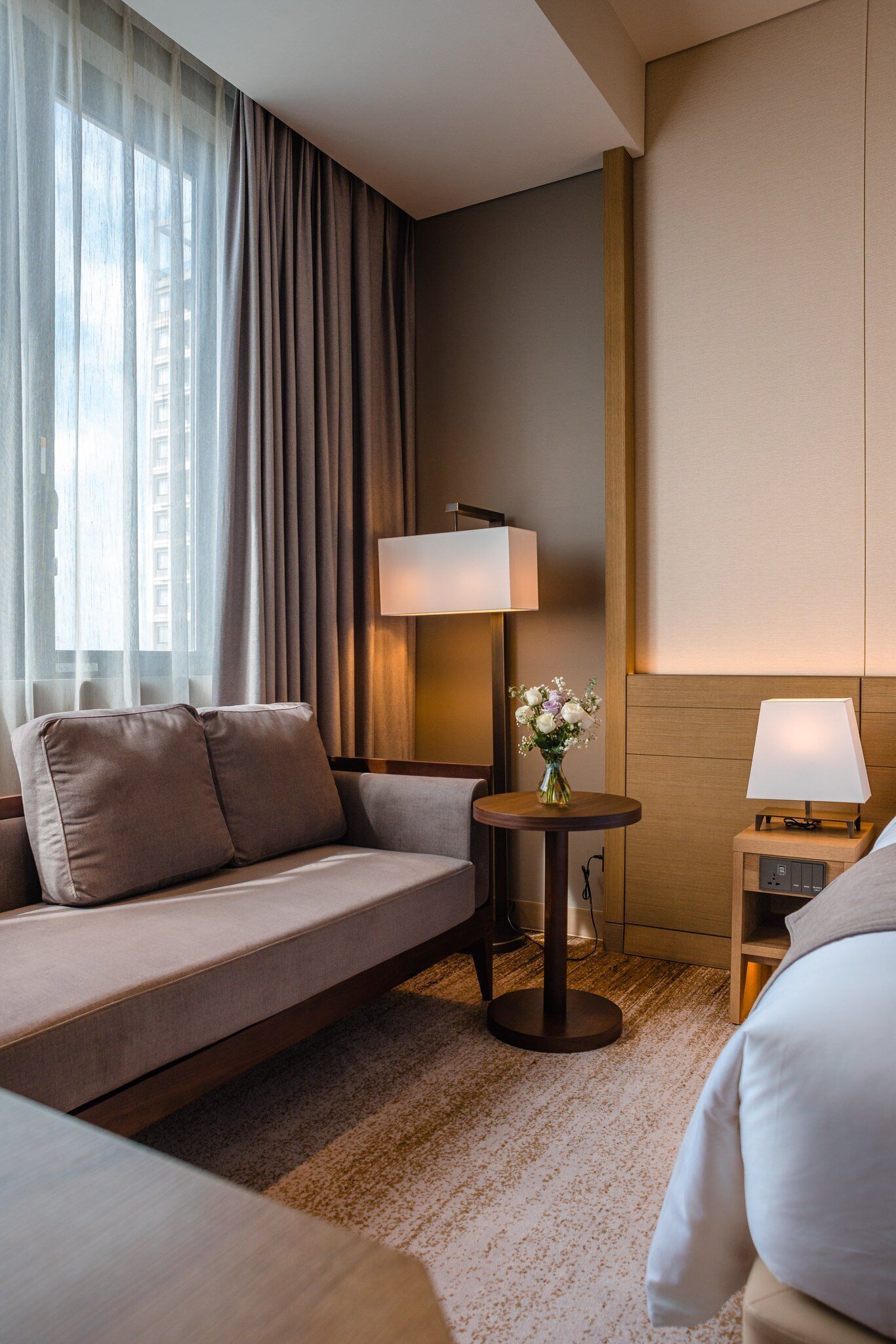 Habitación de hotel con sofá, dos mesitas de noche con lámparas, un pequeño jarrón con flores y vistas a la ciudad a través de cortinas de gasa.