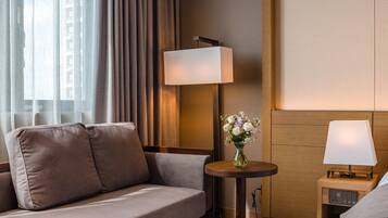 Habitación de hotel con sofá, dos mesitas de noche con lámparas, un pequeño jarrón con flores y vistas a la ciudad a través de cortinas de gasa.
