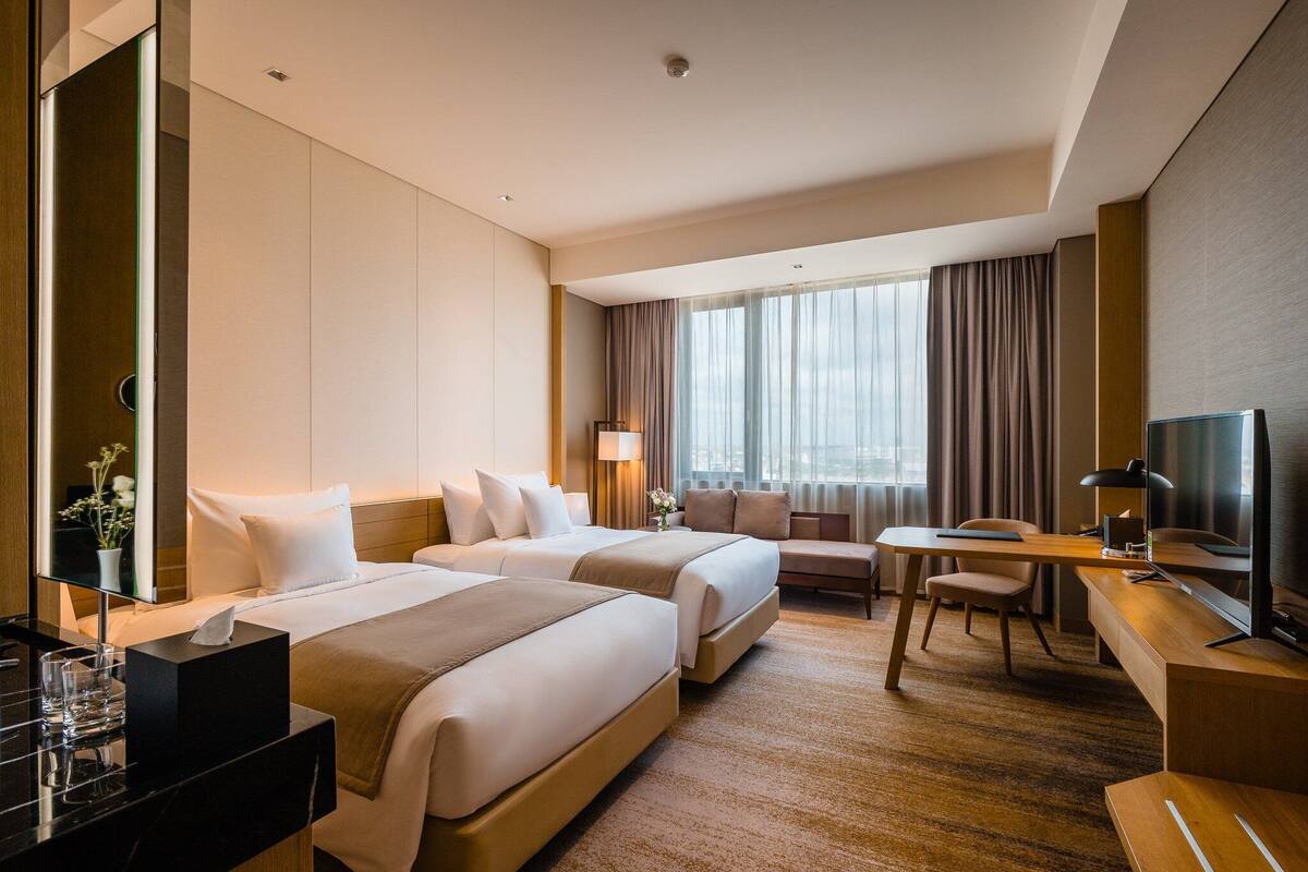 Ein Hotelzimmer mit zwei Betten, einem Schreibtisch, einem Stuhl, einem Fernseher und einem großen Fenster mit Vorhängen.