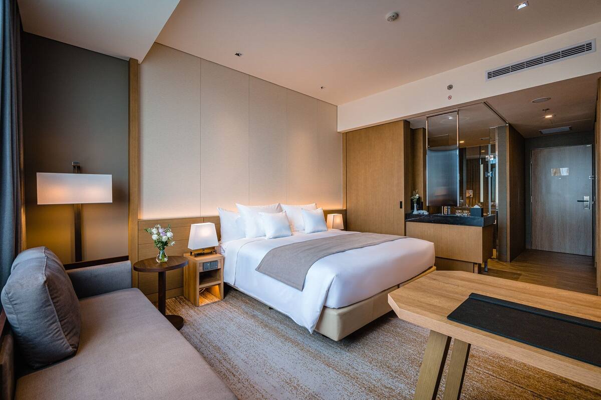 Ein modernes Hotelzimmer mit einem großen Bett, einer Sitzecke, einem Schreibtisch und einem durch eine Glastür einsehbaren Badezimmer.