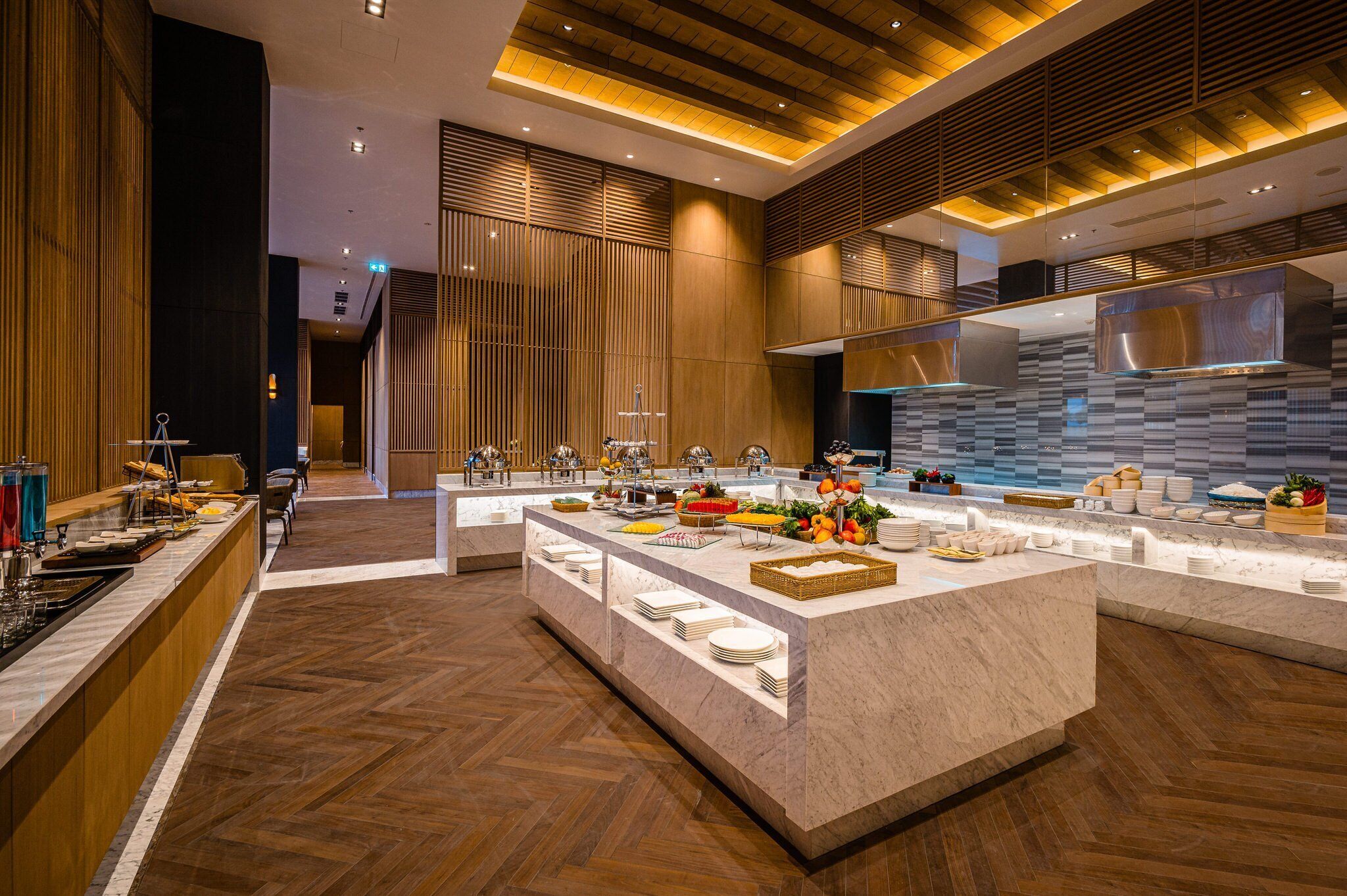 A modern hotel lobby with a central buffet area, wooden paneling, and a herringbone floor pattern.
