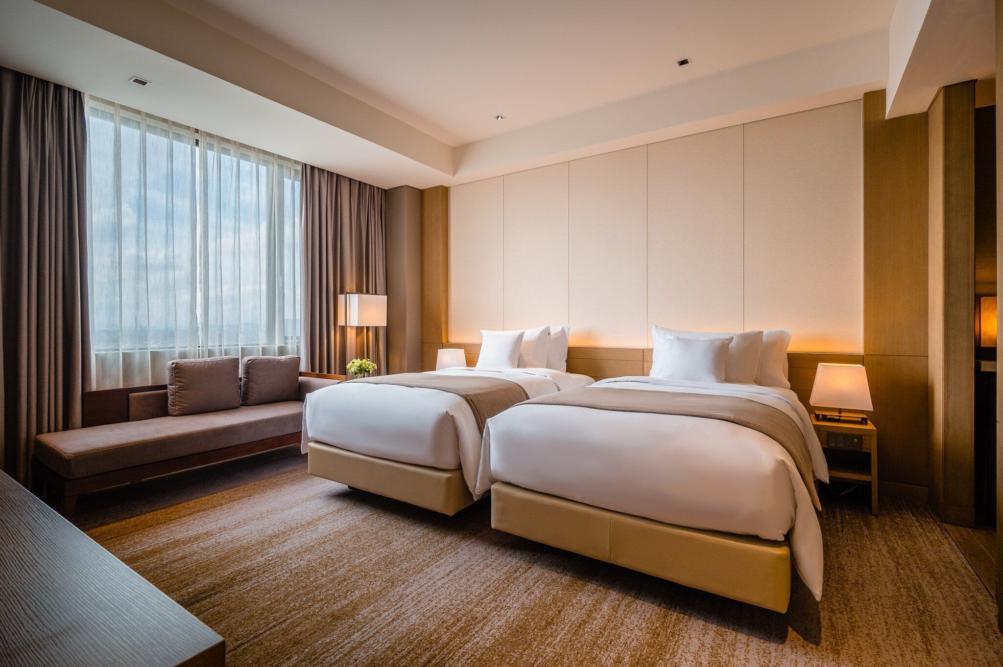 Quarto de hotel com duas camas, um sofá, uma escrivaninha e uma janela grande com cortinas.