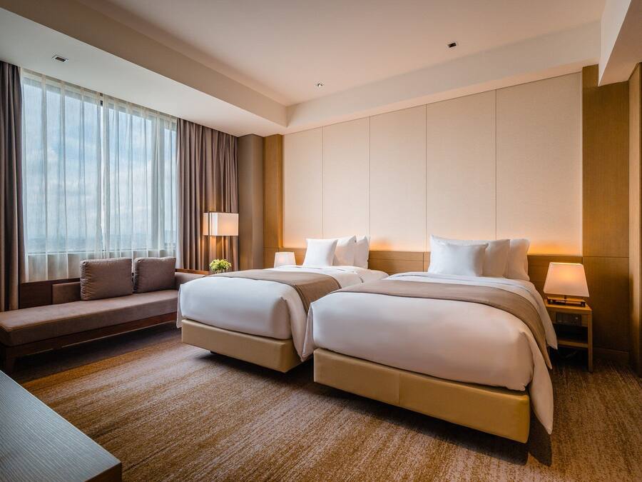 Quarto de hotel com duas camas, um sofá, uma escrivaninha e uma janela grande com cortinas.