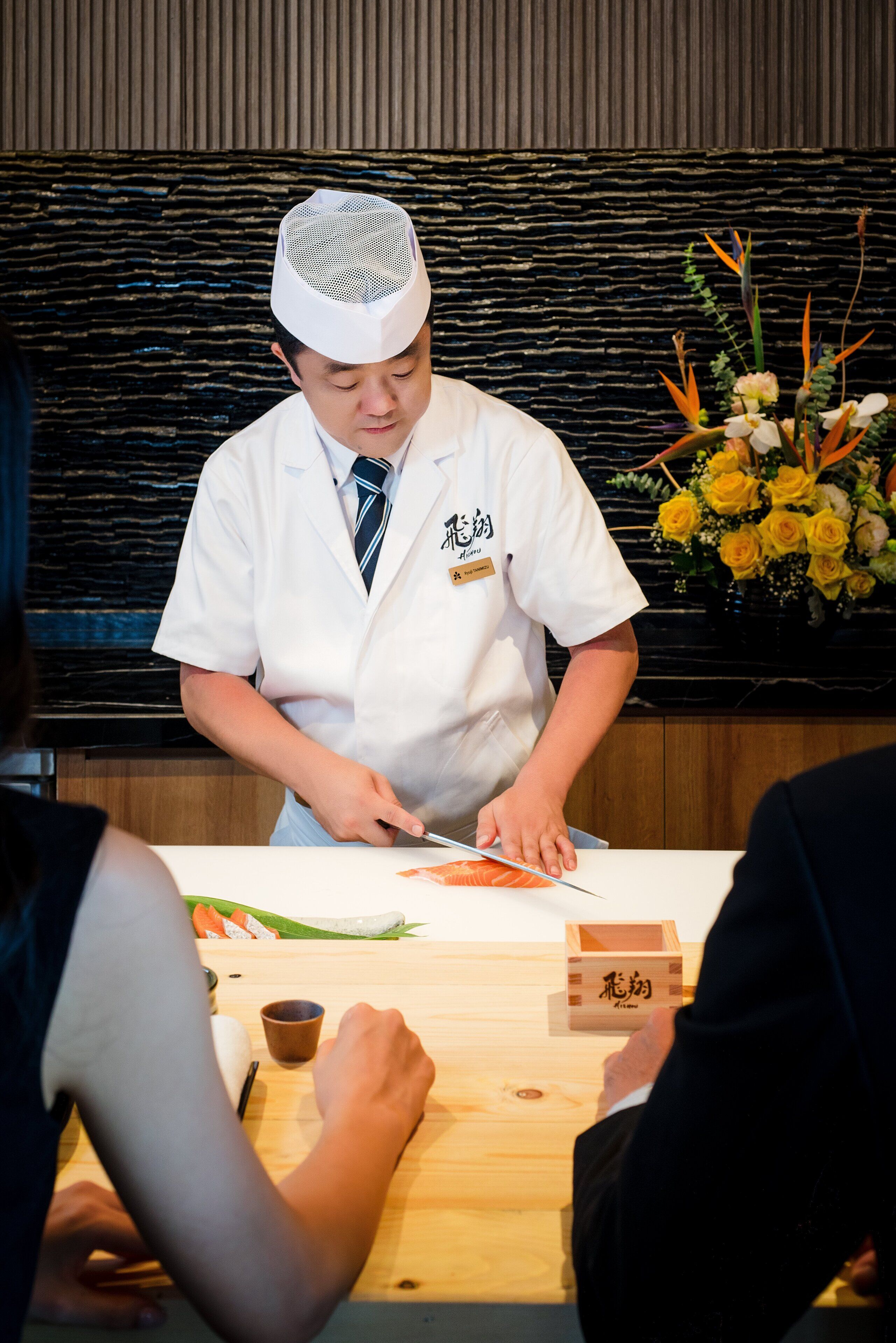 A chef in a white uniform preparing food at a wooden table with a black textured wall in the background.