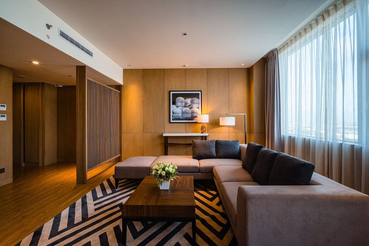 Ein modernes Hotelzimmer mit einer Couch, einem Couchtisch mit Blumen, einer Lampe und einem großen Fenster mit Vorhängen.