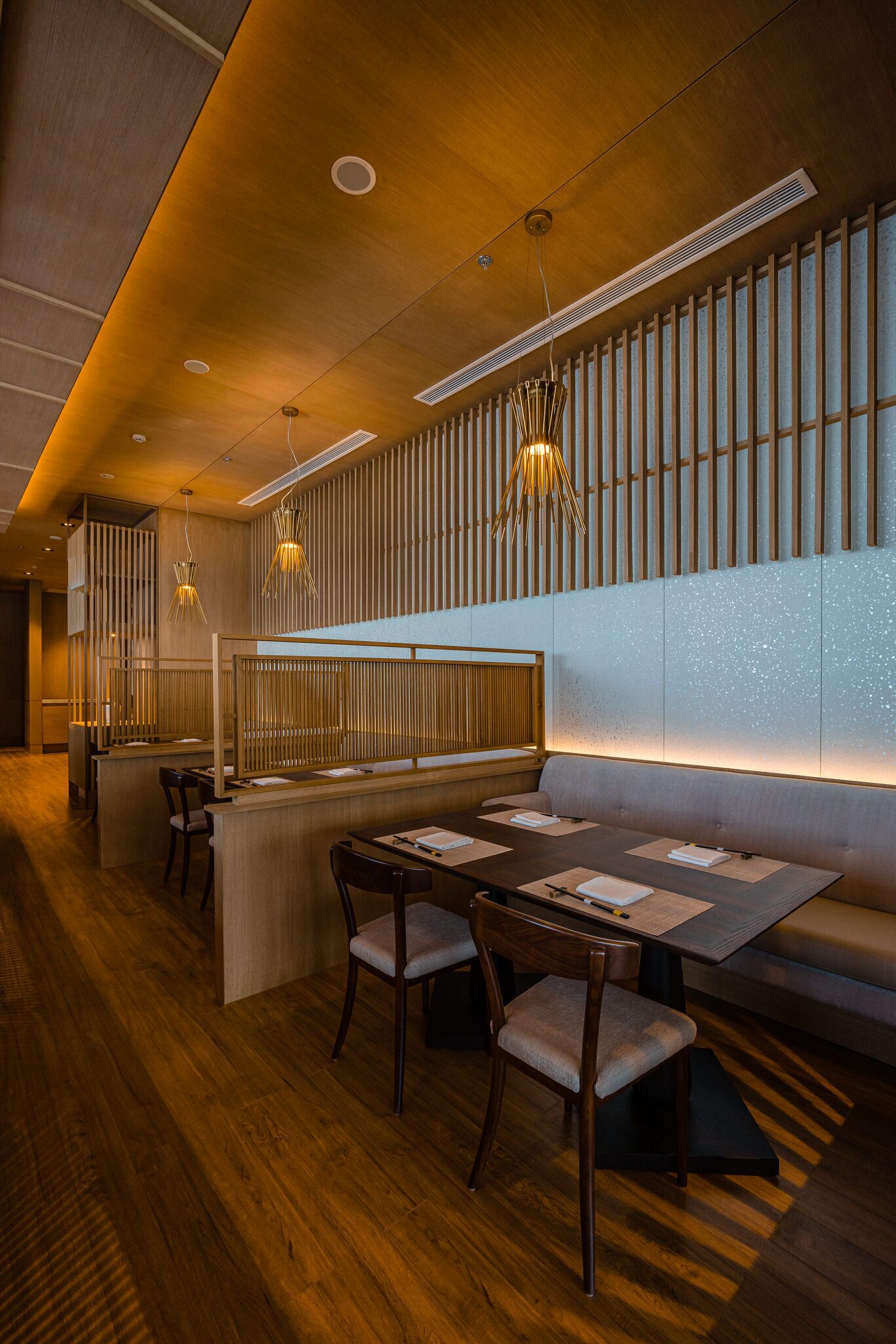 A modern restaurant interior with wooden flooring, a long dining table with chairs, and a bar area with stools.