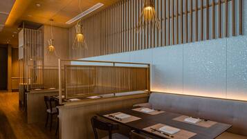 A modern restaurant interior with wooden flooring, a long dining table with chairs, and a bar area with stools.