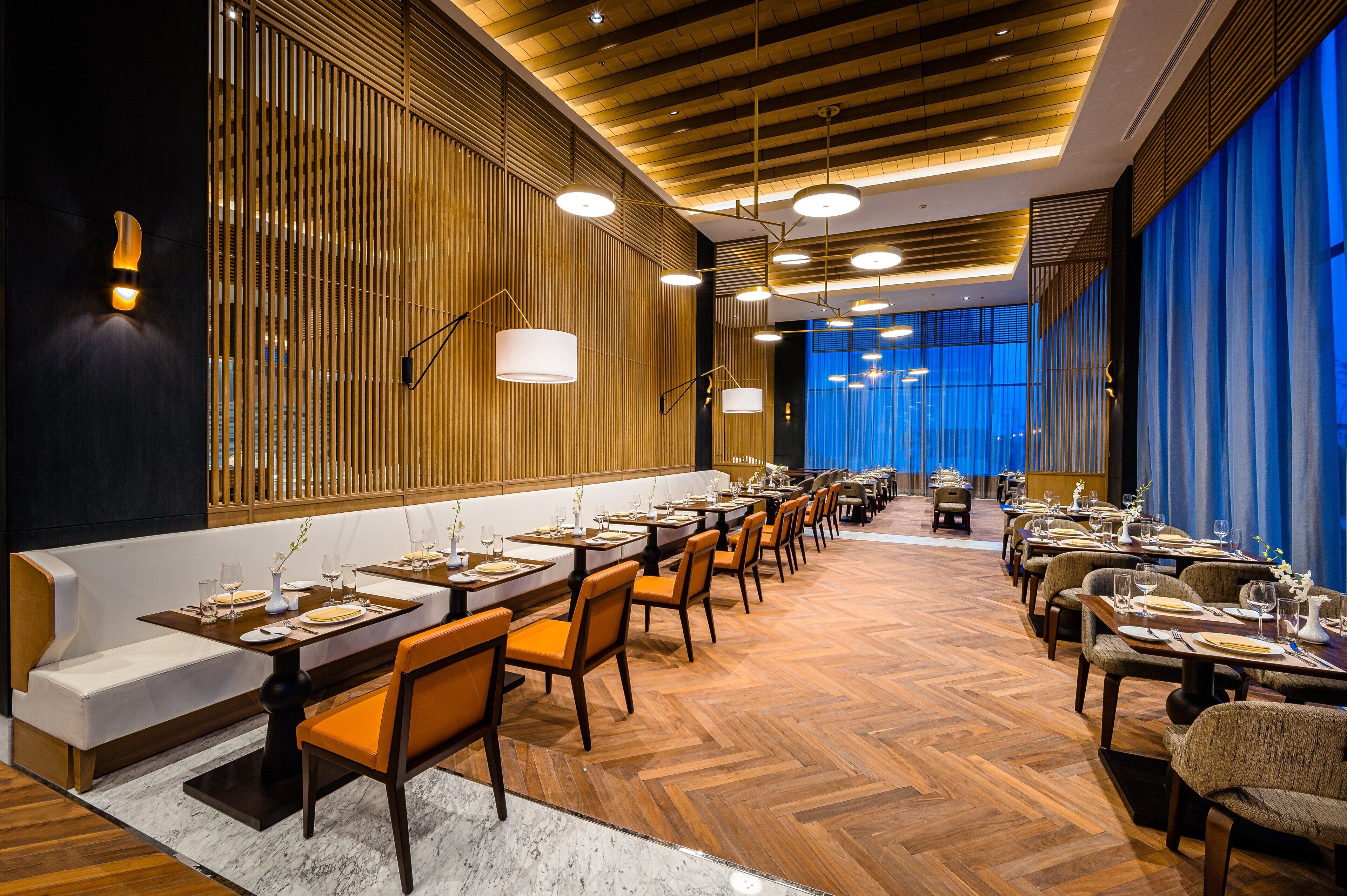 A restaurant with long wooden tables and orange chairs, a marble floor, and a blue curtain backdrop.